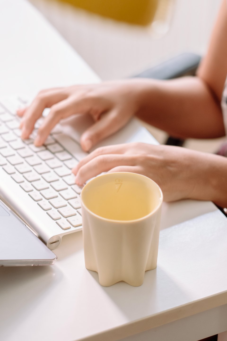 Hände tippen auf einer weißen Tastatur neben einer gelben Tasse.